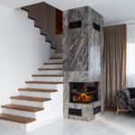 Interior of stylish living room with white stairway and wooden steps placed near marble column with burning fireplace at home